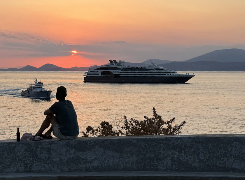 A yacht moored in the bay, sun setting behind it. A young boy sits on a wall in the foreground.