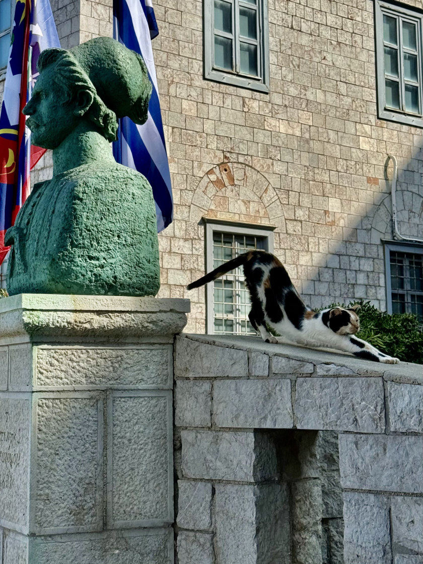 A cat stretching on a wall with a bust of a naval hero to the left.