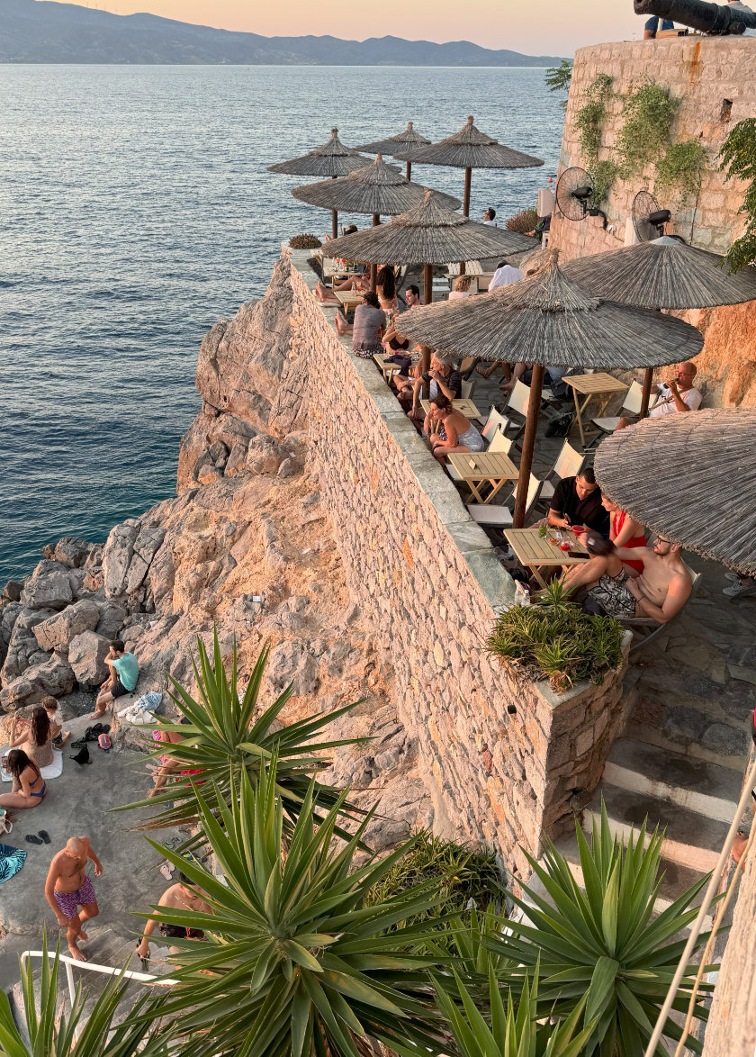 People sitting at tables high above the water, looking out at the view. Others are down below in a flat area amongst the rocks.