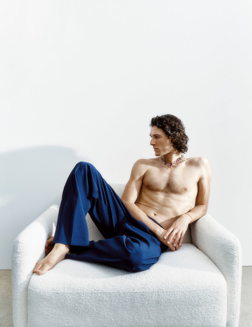 Man posing on chair, topless, wearing necklace